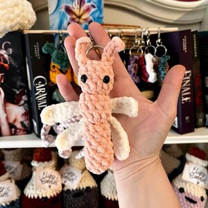 Puede incluir: Un llavero de amigurumi de crochet rosa y blanco con forma de pequeña criatura con ojos grandes. El llavero está siendo sostenido por una mano.