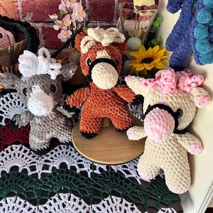 May include: A collection of handmade crochet horse figurines in various colors. The horses have white, pink, and brown manes and muzzles, with black accents. They are displayed on a table with decorative crochet lace and floral accents.