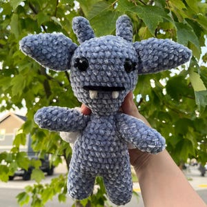 May include: A handmade, plush, blue crochet creature with large ears, wings, and a toothy grin. The toy has black eyes and is held up against a background of green leaves and a bright sky.