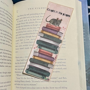 May include: A pink and white bookmark with a mouse sitting on a stack of books. The bookmark has the text "Caught in the pages" on it.