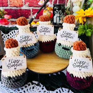 May include: Five crocheted potion bottles with white, gray, green, burgundy, and red colors. Each bottle has a brown cork top and a white tag that says "Elixir Buddies".