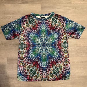 May include: A short-sleeved t-shirt with a vibrant, kaleidoscopic pattern. The design features a symmetrical arrangement of geometric shapes and colors, including blue, green, red, and white. The shirt is made of a soft, lightweight material.