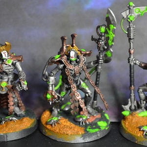 Puede incluir: Tres figuras en miniatura de guerreros robóticos, pintadas en plata, bronce y verde. Cada figura se encuentra sobre una base circular con una superficie texturizada y arenosa. Los guerreros están armados con una guadaña y un bastón.