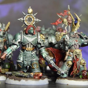 Puede incluir: Un grupo de cinco figuras en miniatura pintadas de verde y rojo, que representan marines espaciales, con armaduras y armas detalladas. Las figuras están en formación de batalla, con una figura sosteniendo un arma con forma de cráneo.