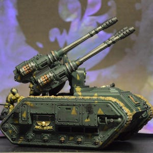 May include: A green and brown military tank model with two large cannons. The tank has a camouflage pattern and a skull and wings symbol on the side.