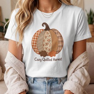 May include: A white short-sleeve t-shirt featuring a large graphic of a pumpkin made of various brown, orange, and cream-colored fabric patterns including plaid, floral, and houndstooth. The text Cozy Quilted Harvest is printed below the pumpkin.