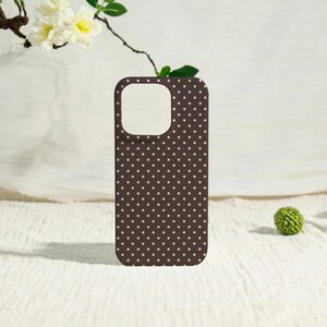 May include: A brown phone case with a repeating pattern of small, light pink polka dots. The case has cutouts for the camera and other phone features. The background is a white, textured surface with a sprig of flowers.
