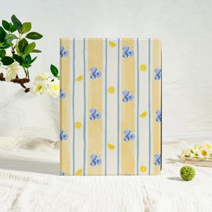 May include: A tablet case with a yellow and white striped pattern. The design includes blue blueberries and yellow lemon slices. The case is a rectangular shape and is standing upright on a white surface.