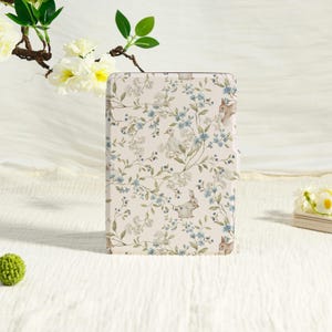 Könnte beinhalten: Eine rechteckige E-Reader-Hülle mit cremefarbenem Hintergrund und einem wiederkehrenden Muster aus kleinen blauen Blumen, grünen Blättern, weißen Blüten sowie Illustrationen von kleinen braunen Kaninchen und einem weißen Entenküken.
