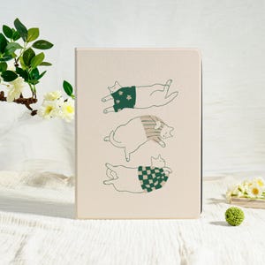 May include: A beige notebook featuring three cartoon cats in various poses. Each cat wears a different patterned sweater: floral, striped, and checkered. The notebook is rectangular and has a simple, minimalist design.