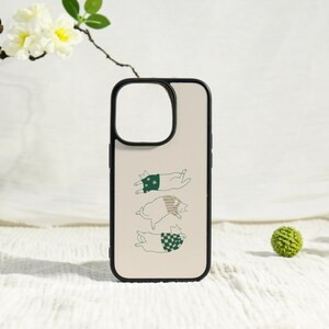 May include: A phone case with a black border and a beige background. The case features a whimsical illustration of three cats wearing sweaters in green and tan. The cats are lying down, creating a playful design.