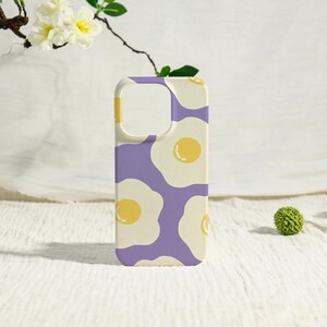 May include: A phone case with a repeating pattern of fried eggs on a purple background. The eggs have yellow yolks and white egg whites. The case has a cream-colored border and a cutout for the camera.
