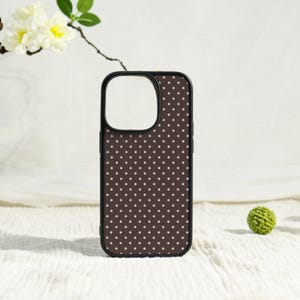 May include: A black phone case with a brown polka dot pattern. The case has a black border and a cutout for the camera. The background is a white surface with a sprig of flowers and a small green ball.