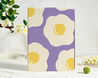 May include: A rectangular notebook featuring a purple backdrop adorned with a pattern of fried eggs. The eggs are white with yellow yolks. The notebook rests on a white surface.