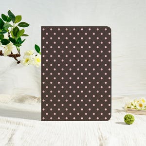 May include: A brown notebook featuring a pattern of small, evenly spaced pink polka dots. The notebook is rectangular and appears to be closed. The background is a soft white.