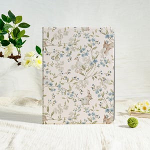 Peut inclure: Un étui rectangulaire avec un motif floral composé de fleurs bleu clair, de feuilles vertes et de petits lapins et canards blancs. L'étui a un fond crème et un bord noir. Le design a une esthétique vintage et printanière.