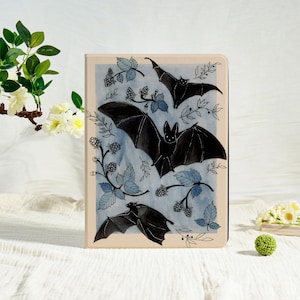 Könnte beinhalten: Eine beige Tablet-Hülle mit einem Aquarelldesign von schwarzen Fledermäusen mit ausgebreiteten Flügeln. Die Fledermäuse sind von blauen Blättern und Beeren auf hellblauem Hintergrund umgeben. Die Hülle hat einen dunklen Rand.