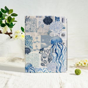Könnte beinhalten: Ein rechteckiges Notizbuch mit einem Collage-Design mit Muscheln, Quallen und Surfbrettern in Blau- und Weißtönen. Das Notizbuch wird auf einer weißen Oberfläche mit floralen Akzenten präsentiert.