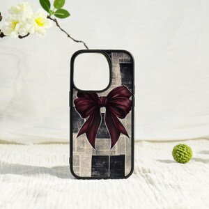 May include: A black-bordered phone case featuring a central dark burgundy bow graphic set against a background of black and white newspaper print. The case has a large square cutout for the camera lens and a sleek, rectangular protective design.