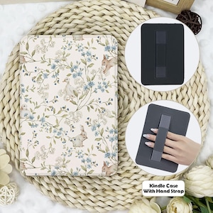 Könnte beinhalten: Eine rosa-weiße Kindle-Hülle mit floralem Muster und einem schwarzen Handgelenkband. Die Hülle ist auf einer geflochtenen Strohoberfläche abgebildet. Der Text "Kindle Case With Hand Strap" ist unter der Hülle gedruckt.