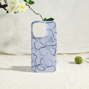 May include: A light purple phone case featuring a floral pattern in a darker shade of purple. The case has a rectangular shape with cutouts for the camera and buttons. The background is a soft, white fabric.