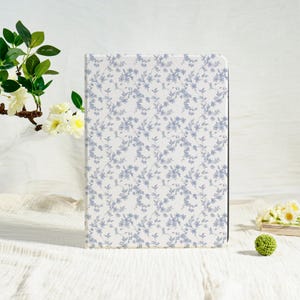 May include: A rectangular notebook with a white background and a delicate blue floral pattern. The cover features a repeating design of small blue flowers and leaves. The notebook is standing upright, suggesting it is new and unused.