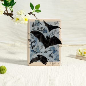 May include: A rectangular e-reader case with a light beige frame featuring a central illustration of three black bats flying amidst blue watercolour-style blackberry vines, leaves, and berries against a soft, pale blue background.