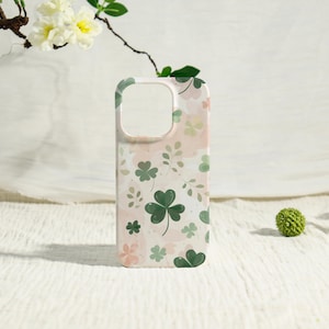 May include: A phone case with a St. Patrick's Day theme. The case features a pattern of green shamrocks, pink flowers, and green leaves on a white and pink background. The case has a camera cutout and a smooth finish.
