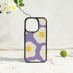 May include: A black phone case featuring a repeating pattern of fried eggs on a purple background. The eggs have yellow yolks and off-white whites. The case has a black border and a cutout for the camera.