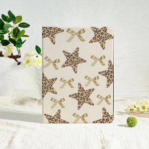 Könnte beinhalten: Ein rechteckiges Notizbuch mit cremefarbenem Hintergrund. Es zeigt ein Muster aus Leopardenmuster-Sternen und goldenen Schleifen. Das Cover ist mit mehreren Sternen und Schleifen verziert, was ein verspieltes und stilvolles Design ergibt.