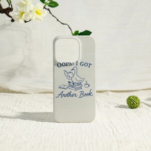 May include: A white phone case featuring a blue graphic of a duck sitting on a stack of books while reading, accompanied by a cup of coffee and the text OOPS! I GOT Another Book printed in a dark blue font above and below the illustration.