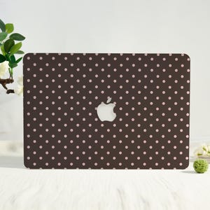 May include: A brown laptop case with a pattern of small, evenly spaced pink polka dots. The Apple logo is visible in the center. The case is designed to fit a laptop and provides a decorative and protective cover.
