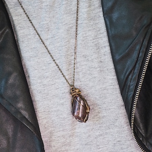 Op de afbeelding: Een zilveren ketting met een hanger van donkerpaarse steen gewikkeld in koperdraad.