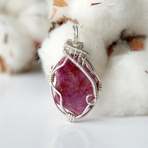 May include: A handcrafted pendant featuring a vibrant, oval-shaped, ruby gemstone. The ruby is encased in a delicate, silver-colored wire wrap, creating a unique and elegant design. The pendant hangs from a small loop at the top.