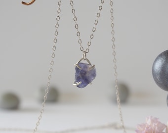 May include: A delicate silver necklace featuring a raw, irregular-shaped, blue-purple gemstone pendant. The pendant is held in a silver setting and hangs from a fine silver chain. The background is blurred, with soft, neutral tones.