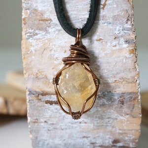 May include: A copper wire wrapped pendant featuring a yellow citrine gemstone. The pendant is attached to a black leather cord.