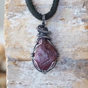 May include: A pink ruby gemstone pendant wrapped in silver wire, hanging from a black leather cord.