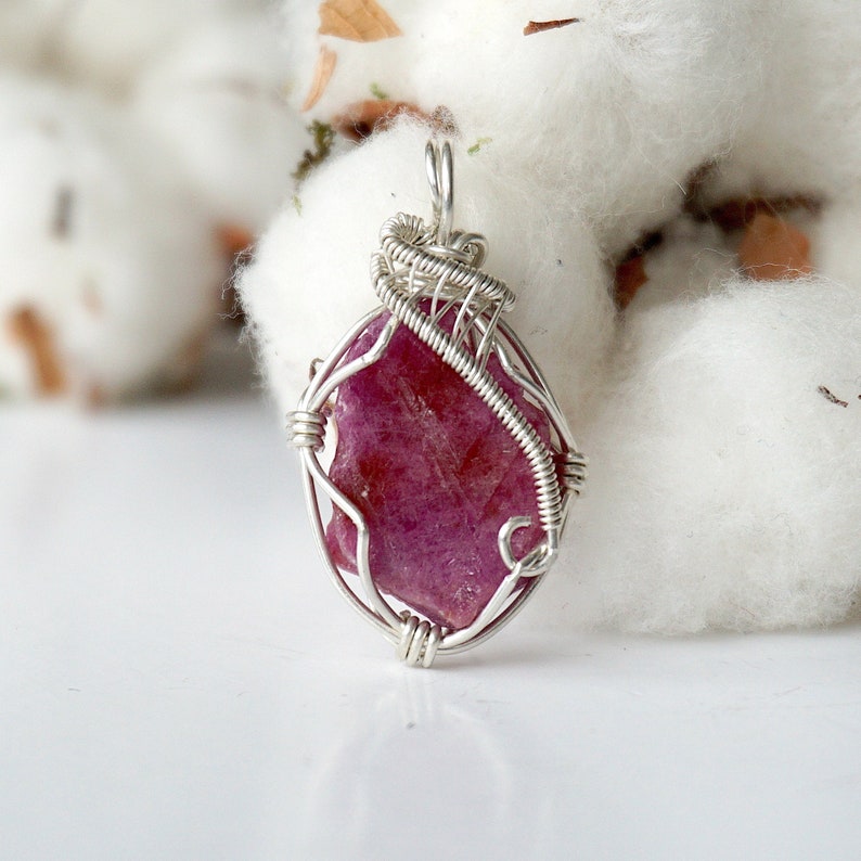May include: A raw, deep pinkish-red gemstone pendant secured in a silver-colored wire-wrapped setting. The stone has an irregular, natural shape and is held by thin metal wires that form a decorative cage around the perimeter, ending in a loop.