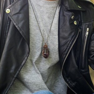 May include: A dark, reddish-purple gemstone pendant necklace. The stone is wrapped in copper wire and hangs from a delicate chain. The necklace is worn over a gray shirt and against a black leather jacket.