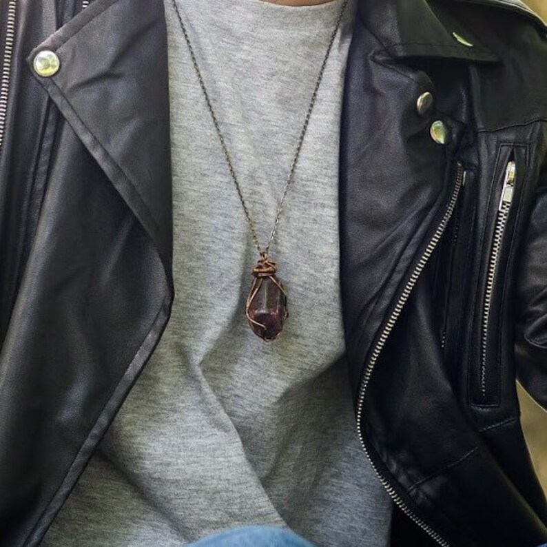 May include: A dark, reddish-purple gemstone pendant necklace. The stone is wrapped in copper wire and hangs from a delicate chain. The necklace is worn over a gray shirt and against a black leather jacket.