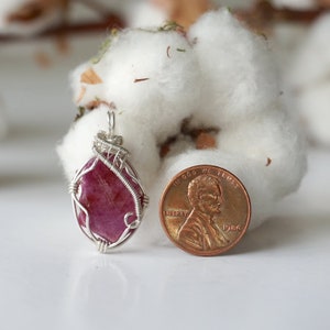 May include: A raw, deep pink ruby gemstone pendant encased in a silver-colored wire wrap. The pendant is positioned next to a copper-colored United States one-cent coin for scale, showing the relative size of the stone and its metal setting.