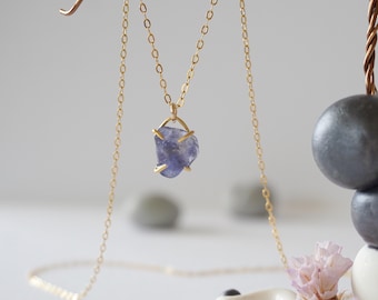 May include: A gold-colored chain necklace with a pendant featuring a rough-cut, blue-purple gemstone. The necklace is displayed against a soft, neutral background, highlighting the jewelry's delicate design and color.