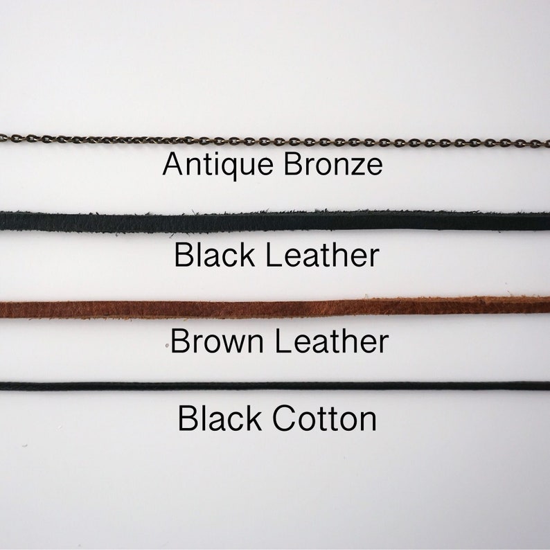 May include: An image showcasing various necklace cord options. The choices include an antique bronze chain, black leather, brown leather, and black cotton cord. Each cord type is labeled with its respective material.