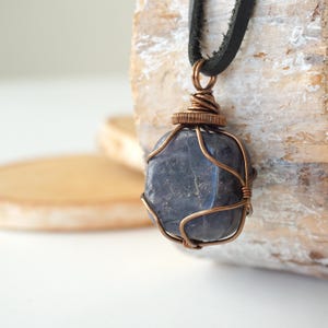 May include: A dark blue, smooth, rounded stone pendant encased in a copper wire wrap. The wire is coiled at the top to form a bail, which is threaded onto a black leather cord. The stone has a polished surface with subtle grey inclusions.