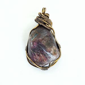 May include: A handcrafted pendant featuring a polished, irregularly shaped stone with shades of purple, brown, and gray. The stone is encased in a wire wrap of gold-colored metal, with a looped design at the top. The pendant is displayed against a white background.