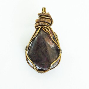 May include: A handcrafted pendant featuring a teardrop-shaped, polished stone in shades of purple and brown. The stone is encased in a gold-colored wire wrap, with a coiled wire detail at the top for hanging. The pendant is set against a white background.