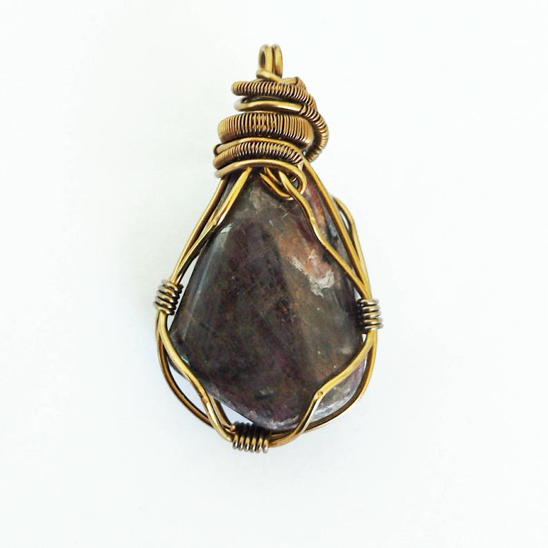 May include: A handcrafted pendant featuring a teardrop-shaped, polished stone in shades of purple and brown. The stone is encased in a gold-colored wire wrap, with a coiled wire detail at the top for hanging. The pendant is set against a white background.