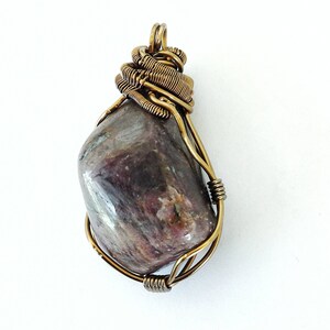 May include: A handcrafted pendant featuring a polished, oval-shaped stone in shades of purple and gray. The stone is encased in a decorative wire wrap of bronze-colored metal, with a loop at the top for a chain. The pendant is set against a white background.