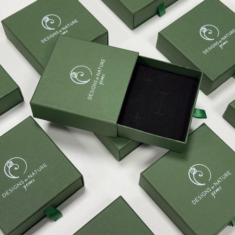 May include: A collection of square, olive green cardboard jewelry boxes with a sliding drawer mechanism. Each box features a white logo with a stylized flower icon and the text DESIGNS BY NATURE gems. One box is open, revealing a black velvet insert.
