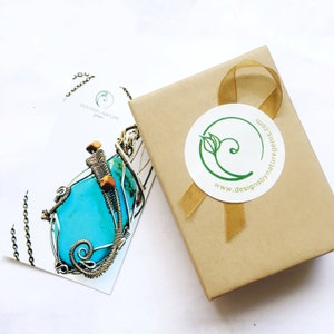 May include: A turquoise gemstone pendant wrapped in silver wire, displayed with a kraft paper gift box tied with a gold ribbon. A card with the logo "designs by nature gems" is also present.